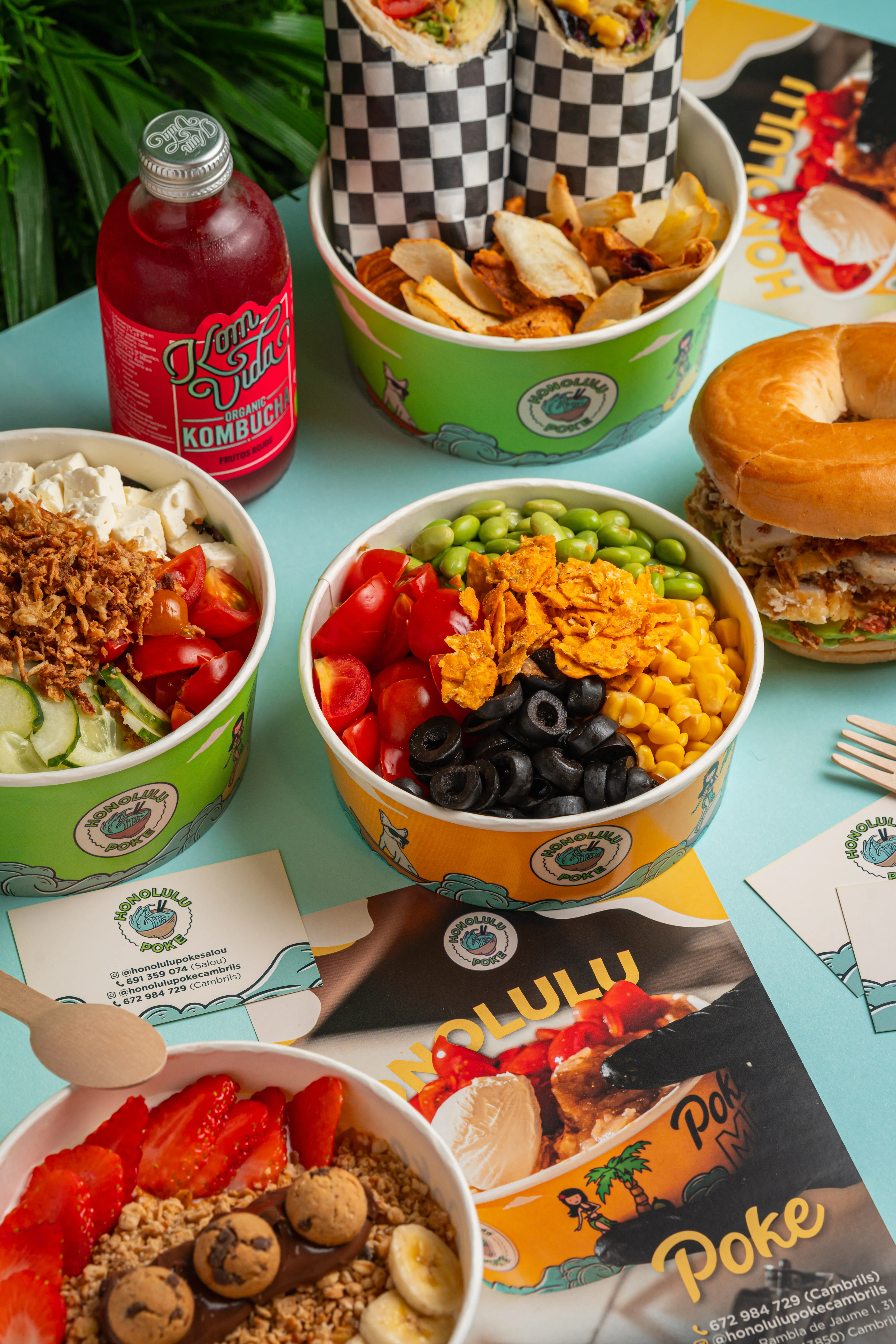 Mesa con varios bowls de Honolulu Poke, wraps, un bagel, una kombucha y tarjetas de contacto de los locales de Salou y Cambrils